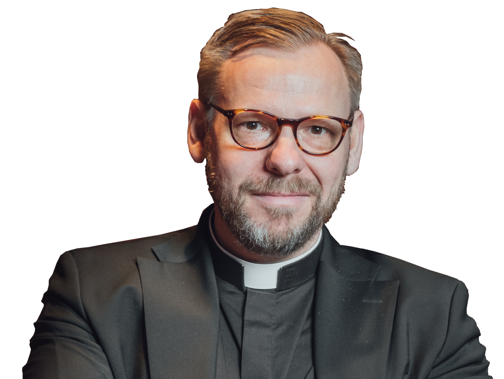 Fred Wilén i klädd svart kostym och prästskjorta. Han har grånande hår och skägg, glasögon och tittar rakt in i kameran.