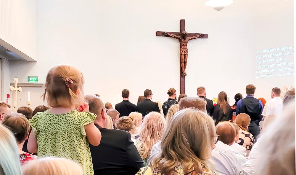 Kuva on otettu konfirmaatiomessusta. Kirkko on täynnä ihmisiä, jotka on kuvattu takarivistä eli heistä näkyy lähinnä selkä ja takaraivo. Kirkkokansan keskellä seisoo tuolilla pieni saparopäinen lapsi vihreässä mekossa. 