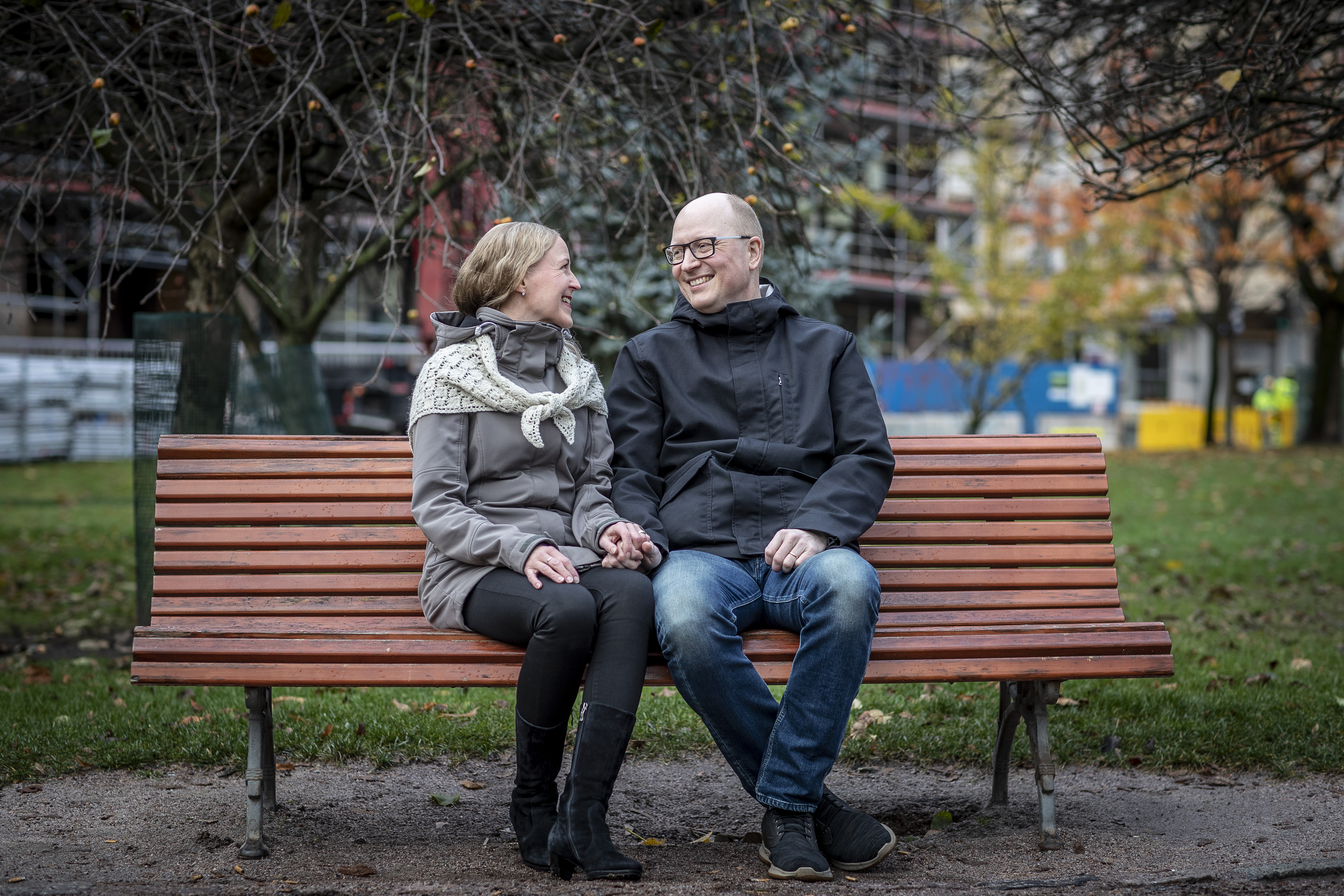 Nainen ja mies istuvat puistonpenkillä. He katsovat toisiinsa ja hymyilevät. Taustalla syksyinen maisema.