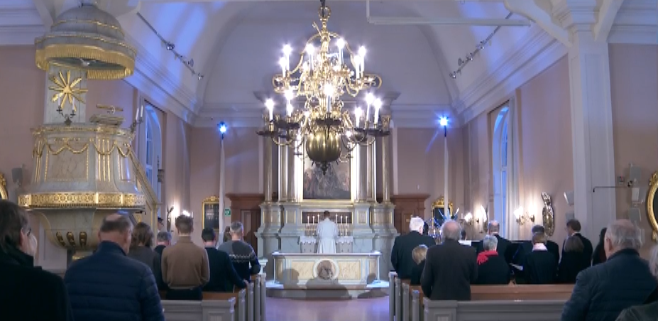 Altaret och en ljuskrona i Jakobstads kyrka är mitt i bild. Till höger och vänster sitter människor i kyrkbänkarna. Framför altaret står prästen klädd i vit alba. Till vänster i bild syns den guldsmyckade predikstolen.