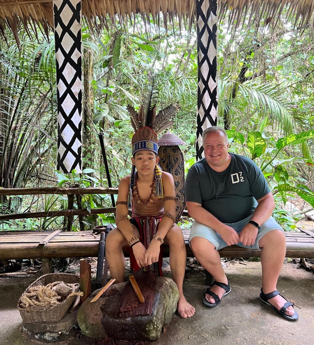 Tomas Ray i kulturcentret Mari-mari i Malaysia. Han sitter till höger i bild i shorts och t-skjorta på en träbänk. Till vänster om honom sitter en malaysier iklädd indianliknande kulturdräkt. I bakgrunden syns djungelväxter och ovanför dem delar av ett basttak.