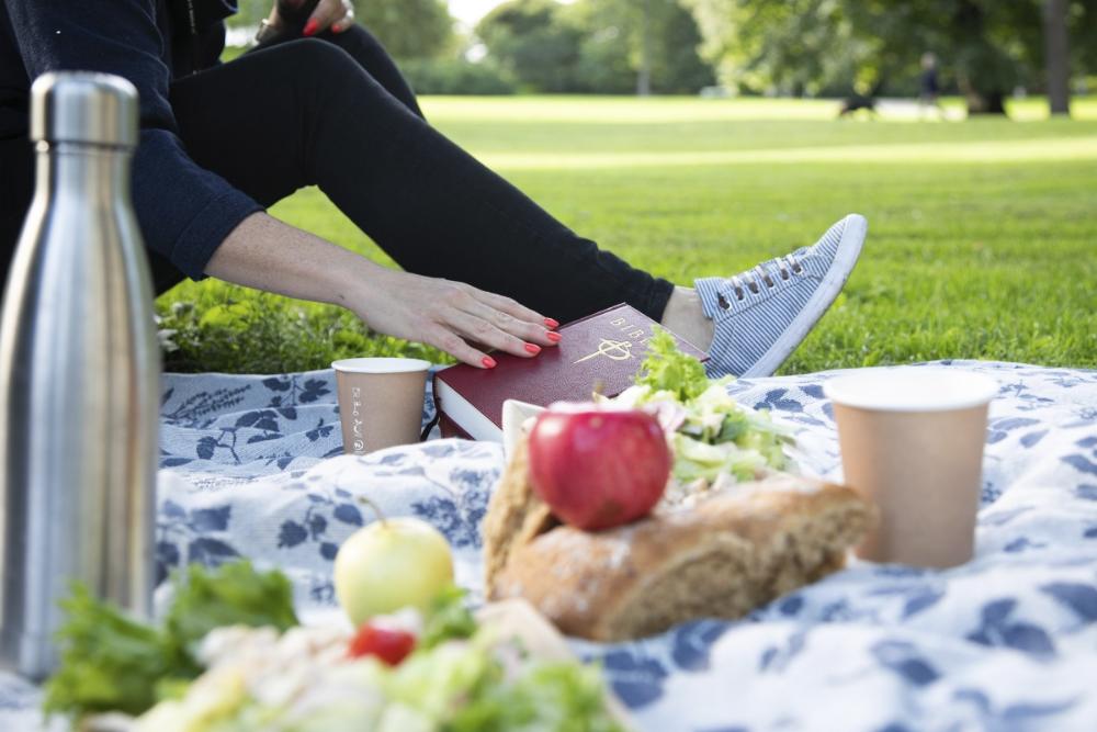 En person sitter på filt på gräsmatta och håller i en bibel. I förgrunden picknick-mat.