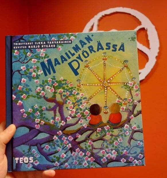 Maailmanpyörässä-kirja kädessä. Kirjan kannessa piirroskuva, jossa kaksi lasta istuu kirsikkapuun oksalla katselemassa maailmanpyörää.