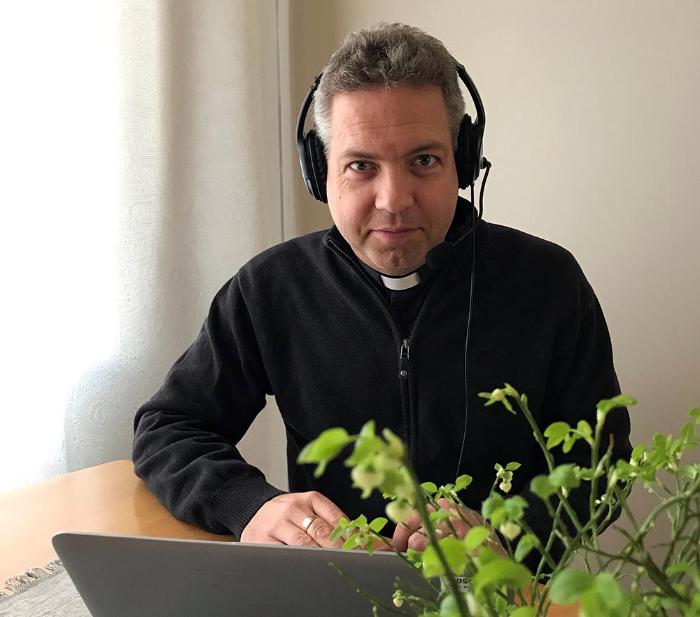 Präst småler i svart prästskjorta med hörlurar framför datorn. Vårljus silar in och björklöv i förgrunden.