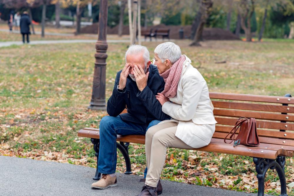 Äldre par sitter på parkbänk, kvinnan tröstar mannen.
