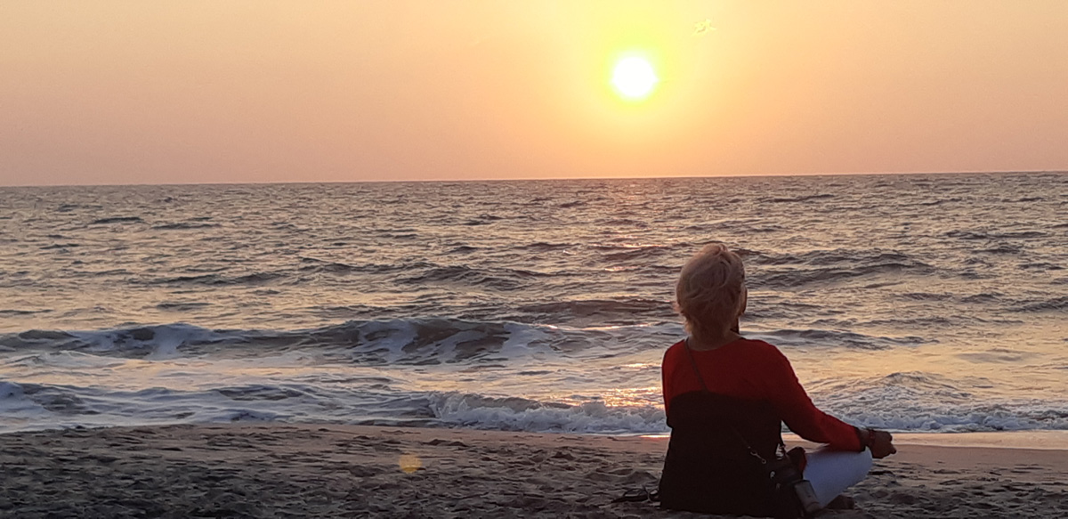 Ihminen istuu rannalla lootusasennossa. Aurinko on lähellä horisonttia.