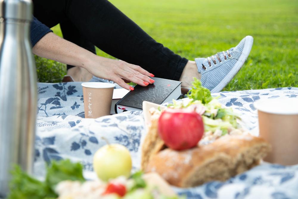 Kesäinen picnic puistossa. Nurmikolla kangas, jonka päällä eväitä. Naisen käsi kurottaa kohti Raamattua.