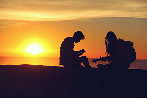 Kaksi nuorta rantakalliolla kauniissa auringonlaskussa kesällä. Toinen soittaa kitaraa.