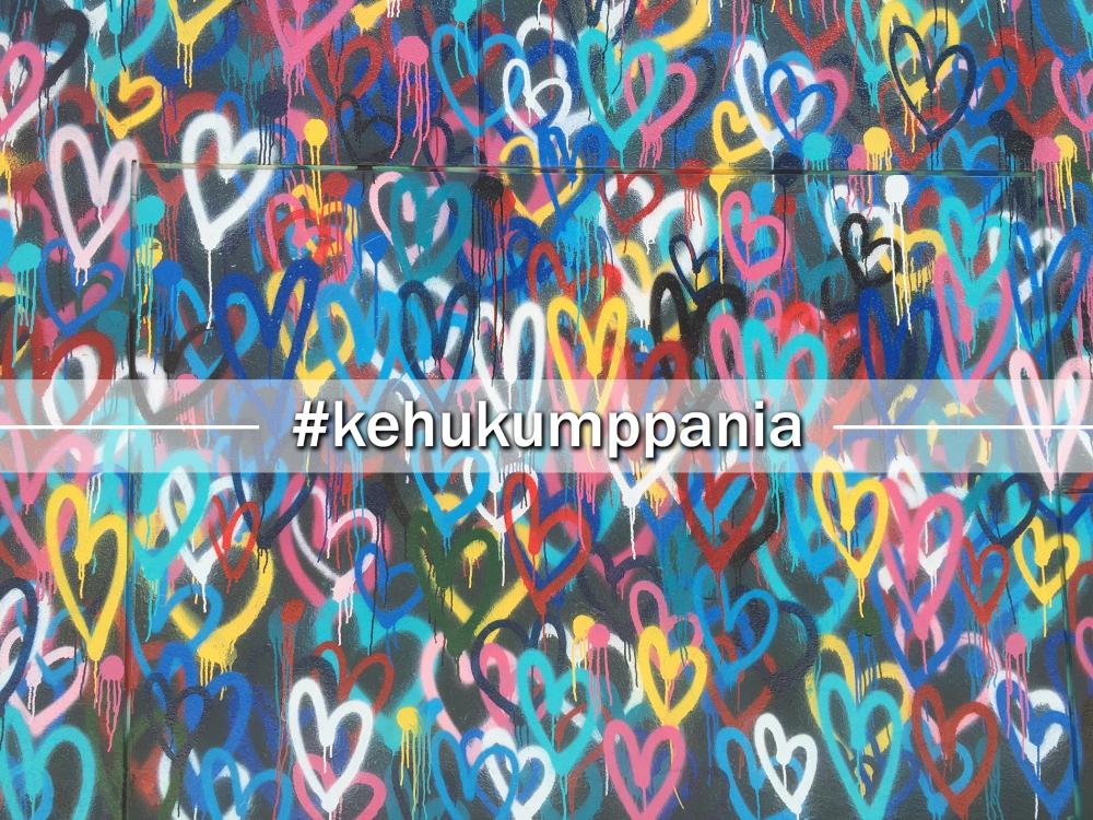 Taustalla seinä maalattuna täyteen värikkäitä sydämiä ja edessä teksti #kehukumppania.