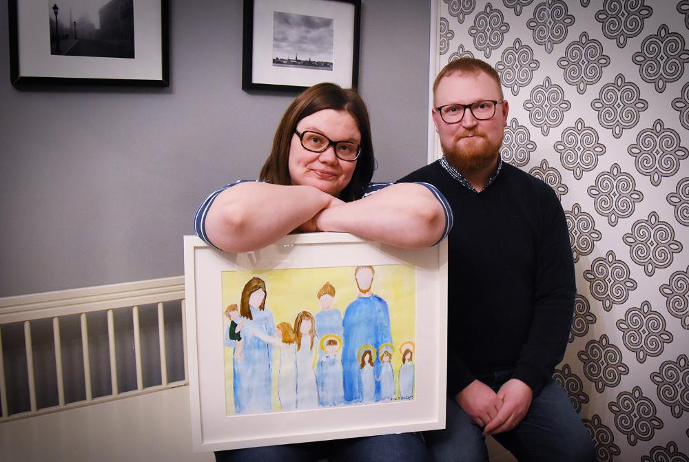 Man och kvinna med en tavla i handen som visar på familjen, där fyra barn är livet och fyra barn har dött.