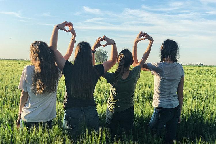 Fyra flickor står på ett fält med ryggen mot kameran och bildar tillsammans hjärtan med sina händer.