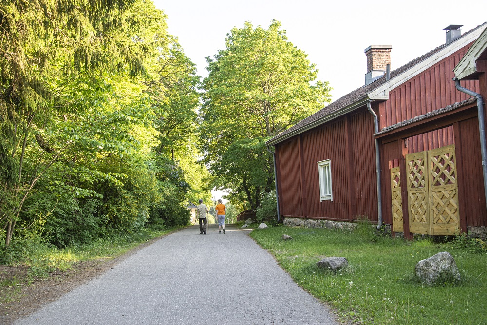 Kaksi vaeltajaa kesäissä iltapäivässä, oikealla punainen puurakennus, vanha tie.
