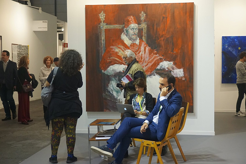 Mies istuu tuolilla ja puhuu puhelimeen taidegalleriassa, jossa on paljon ihmisiä ja maalauksia.
