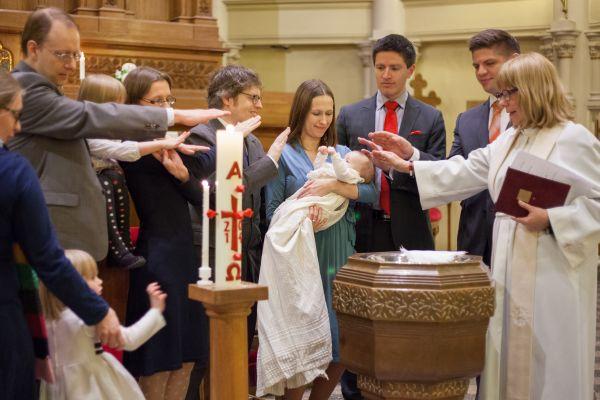 Baby välsignas av präst, familj och faddrar vid dop i kyrka.