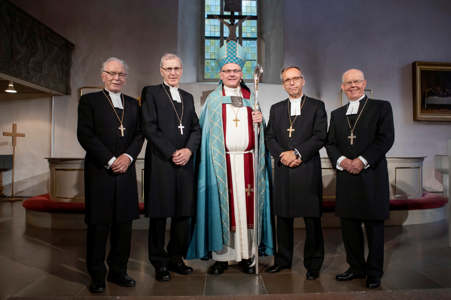 Fem män står uppställda framför altaret i en kyrka. En har ljusblå biskopsdräkt, de andra är svartklädda.