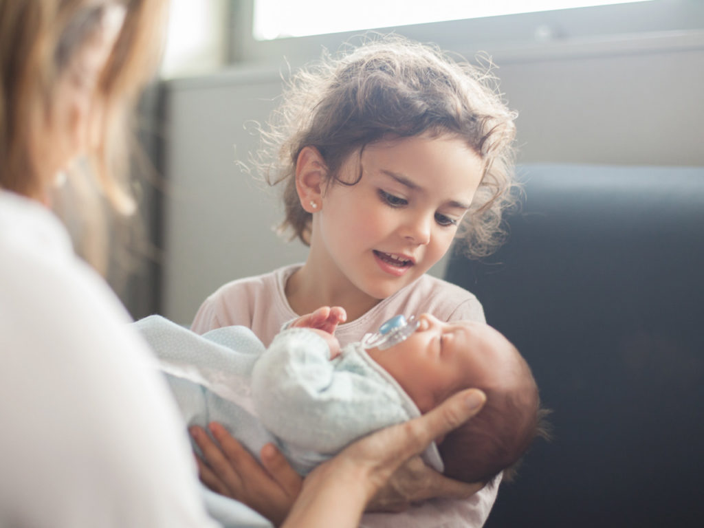 En flicka håller en baby i sin famn med hjälp av en vuxens händer