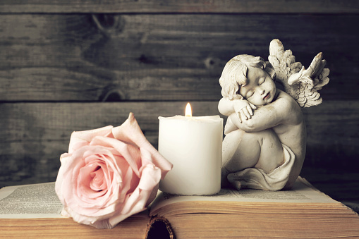 Kuvassa raamatun päällä enkelipatsas, palava kynttilä ja vaaleanpunainen ruusu, puinen seinä taustalla.