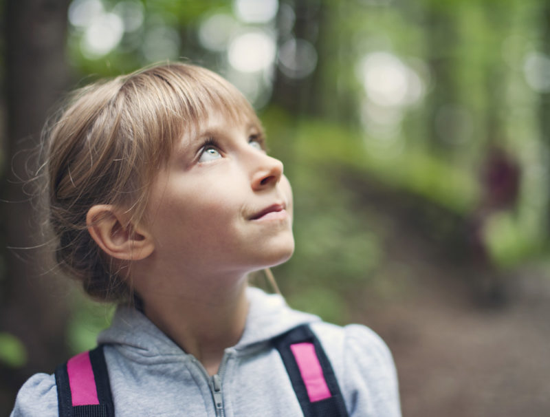 En flicka går på en skogsstig och blickar upp mot himlen.
