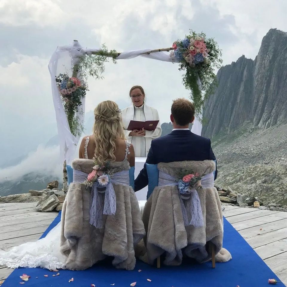Kuvassa hymyilevä pappi toimittaa avioliittoon siunaamisen Sveitsissä vuoristomaisemissa. Pari istuu tuoleilla selkä kameraan päin, katsoen kohti toimittavaa pappia.