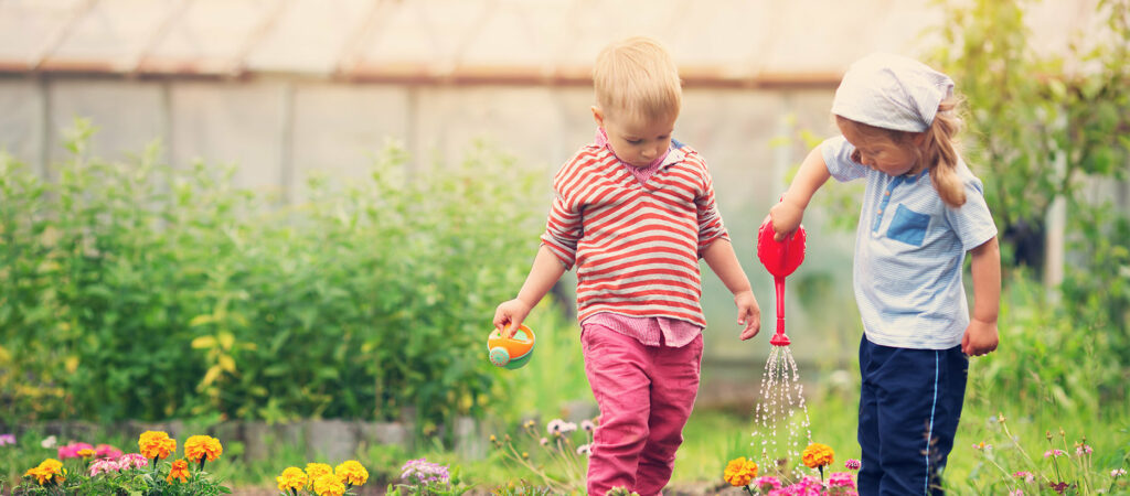 Pieni tyttö kastelee kukkia kastelukannulla ja pikkupoika seuraa vieressä pieni kastelukannu kädessään. 