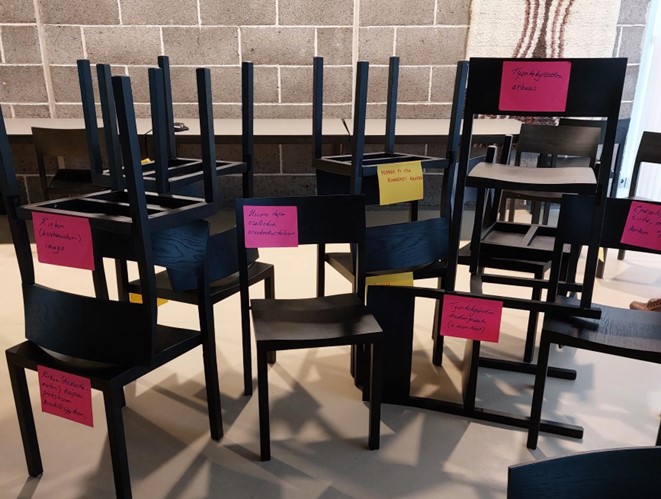 Svarta stolar oorganiserat staplat på varandra som en mur. Stora färggranna post-it-lappar med olika text finns fästa på ryggstöden.