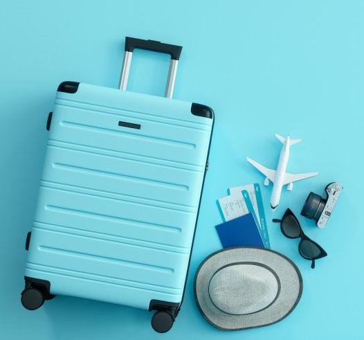 Matkalaukku, hattu, aurinkolasit ja lentokoneen pienoismalli sekä muita matkustamiseen liittyviä pikkutavaroita.