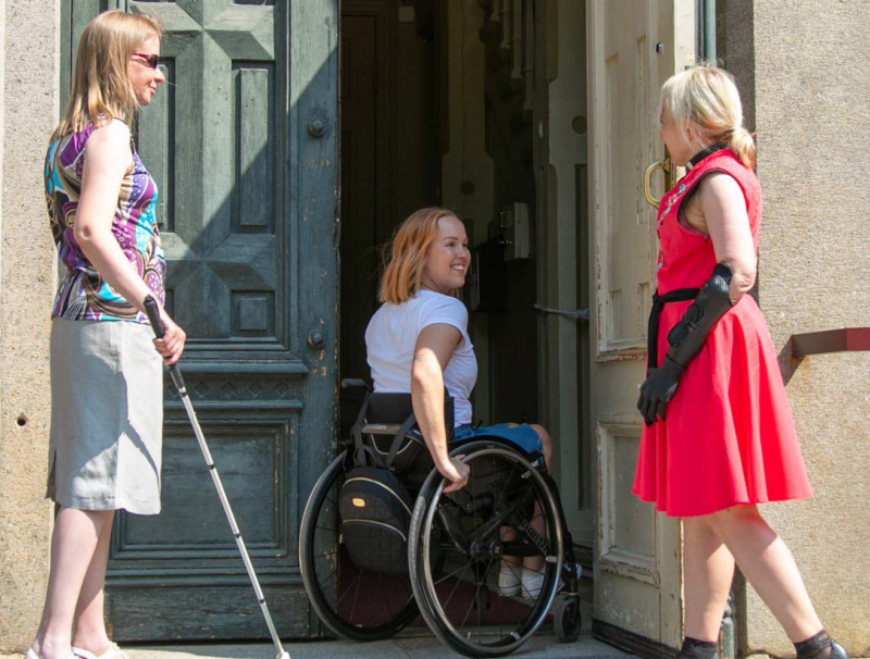 Kolme hymyilevää naista avonaisella kirkon ovella aurinkoisena kesäpäivänä. Yksi käyttää pyörätuolia, toisella on näkövammaisen valkoinen keppi ja kolmannella käsiproteesi.