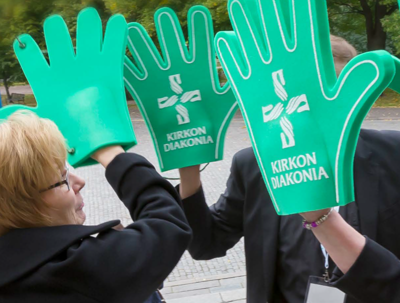 Kolme ihmistä seisoo vastakkain. Jokaisella on kädessään suuri vihreä vaahtomuovikäsi, jossa on Kirkon diakonian logo.