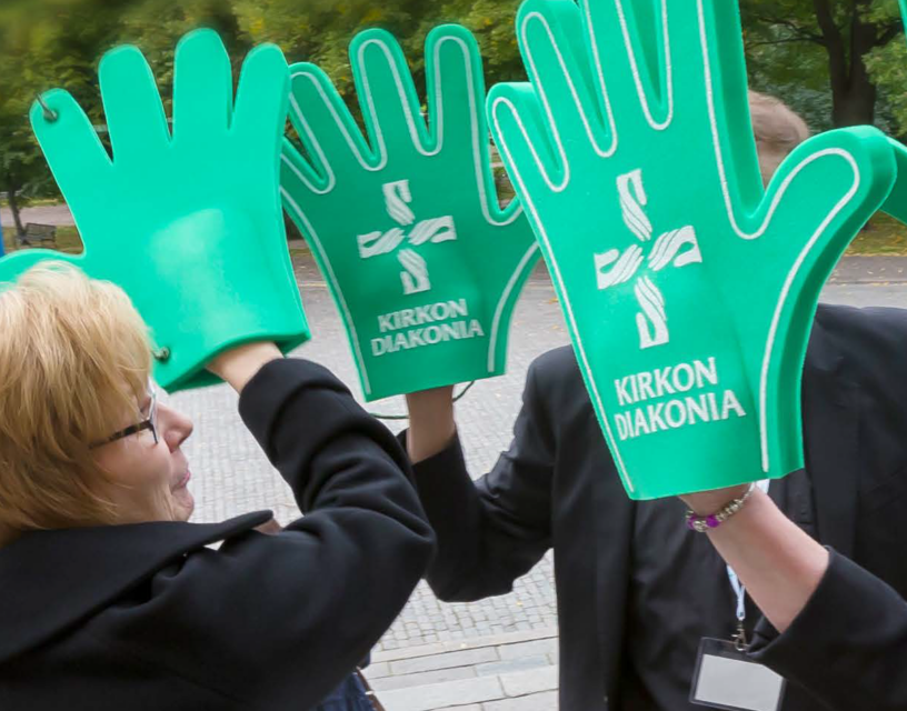Kolme ihmistä seisoo vastakkain. Jokaisella on kädessään suuri vihreä vaahtomuovikäsi, jossa on Kirkon diakonian logo.