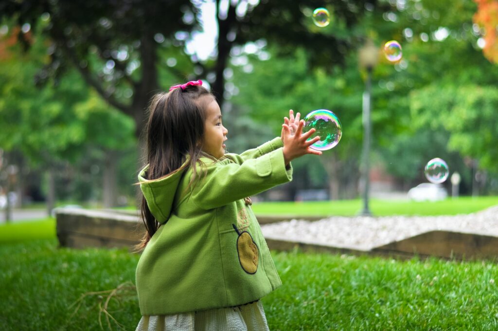 En flicka står i en park och och sträcker händerna mot en såpbubbla.
