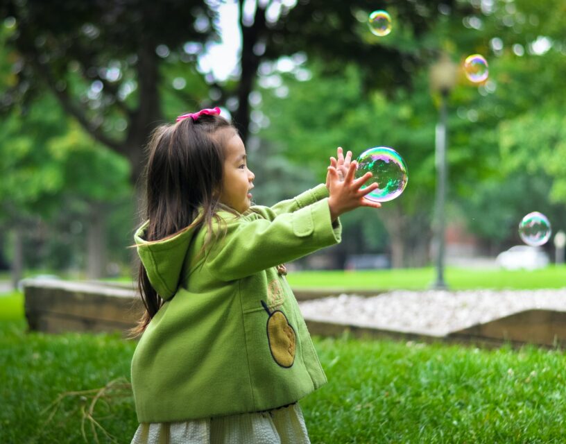 En flicka står i en park och och sträcker händerna mot en såpbubbla.