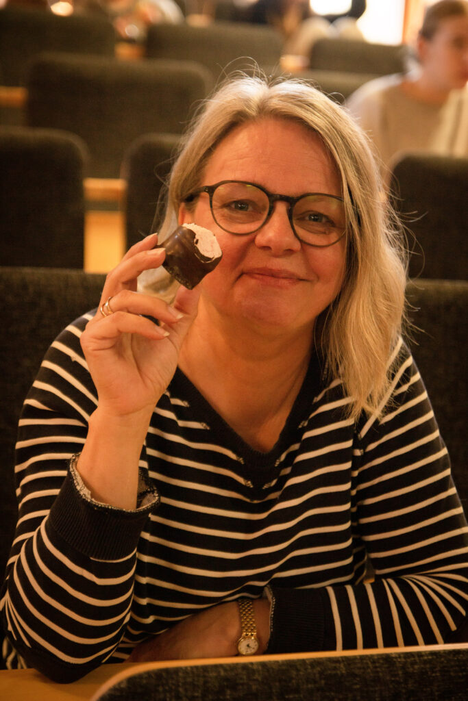 Kvinna i randig skjorta, glasögon och ljust hår håller leende upp en halväten chokladkyss.