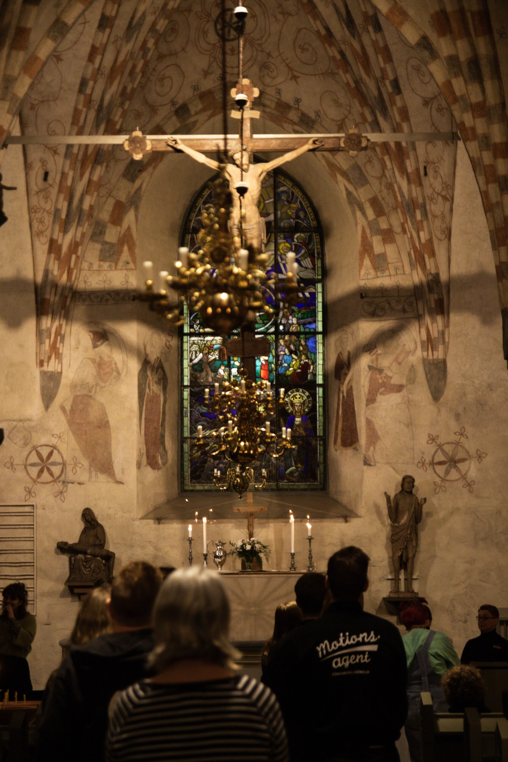 Människor går mot altaret i en medeltida stenkyrka. Ett stort krusifix hänger från taket.