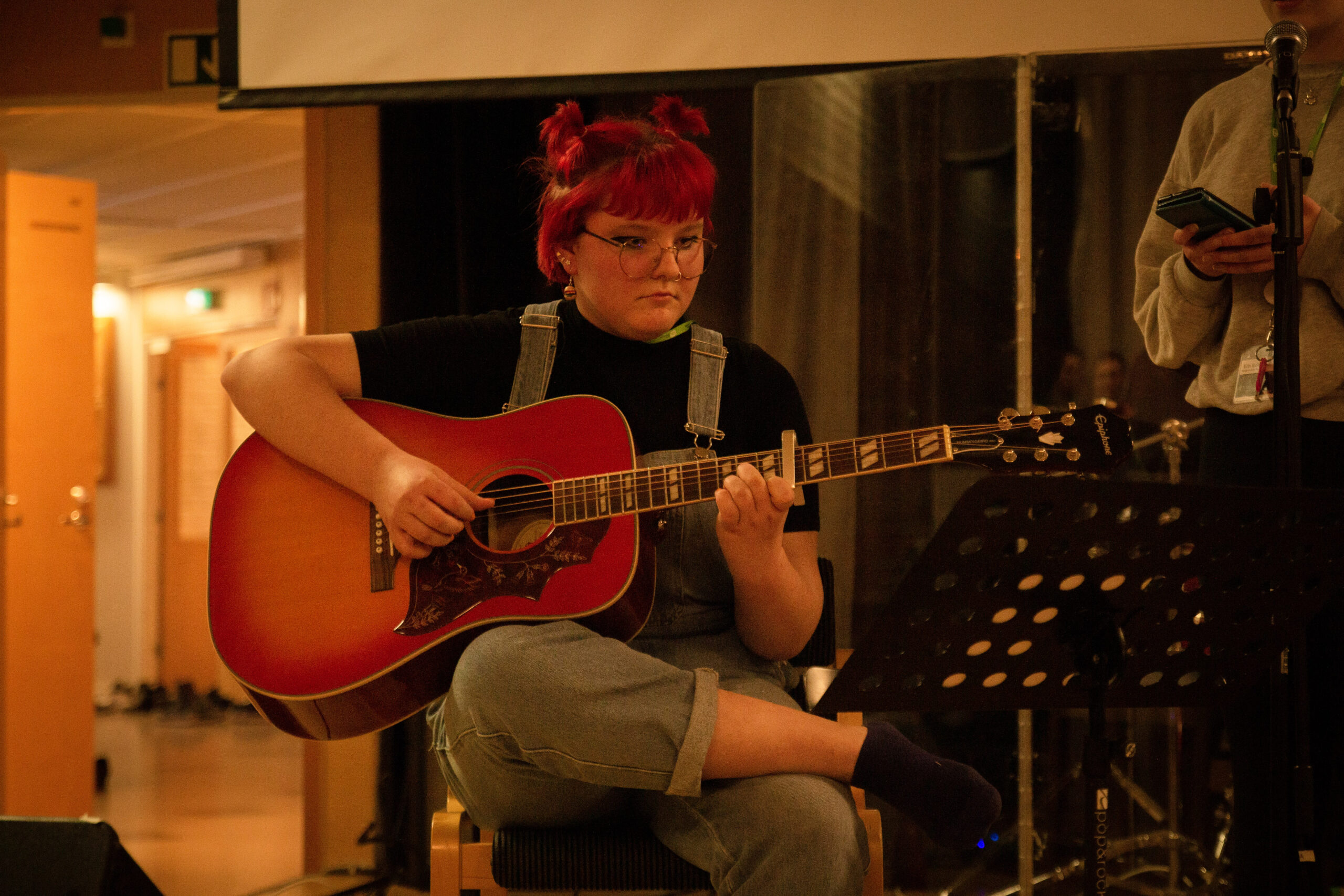 Tonårsflicka med rött hår, glasögon och jeanshalare sitter på stol och spelar gitarr.