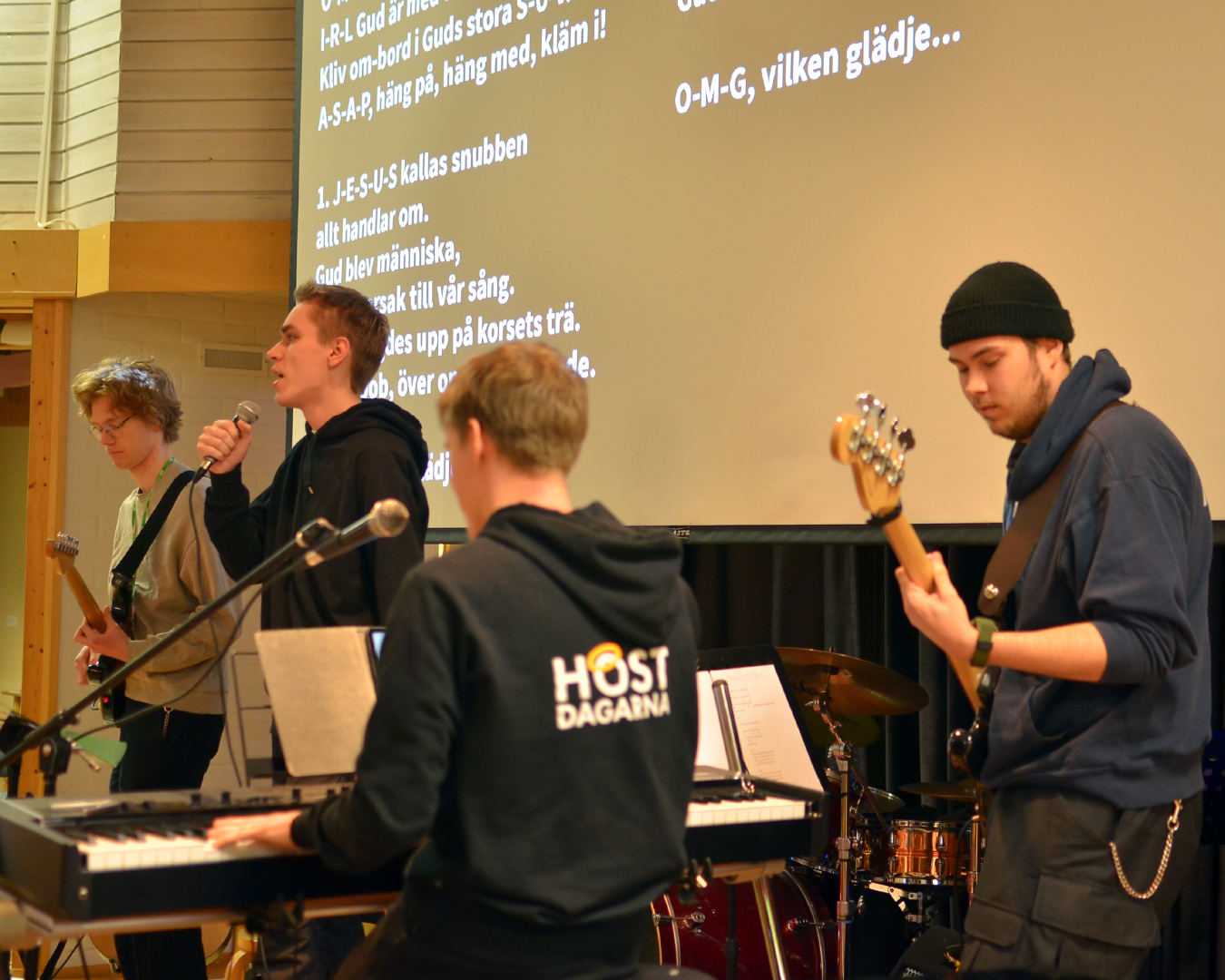 Fyra unga män i 20-års ålder spelar elgitarr, bas, piano och sjunger. Sångtexten syns på en vit skärm i bakgrunden.
