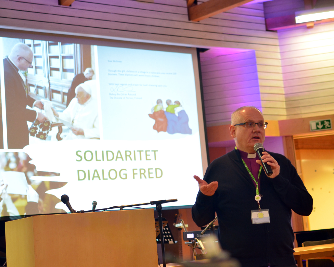 En biskop talar i mikrofon framför en skärm med texten solidaritet, dialog, fred.