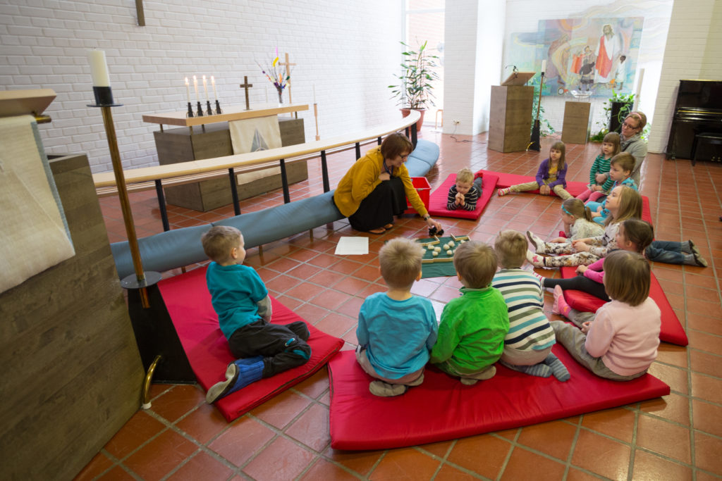 Barn sitter på madrasser vid altaret i en kyrkan och följer med klubbledarens berättelse.