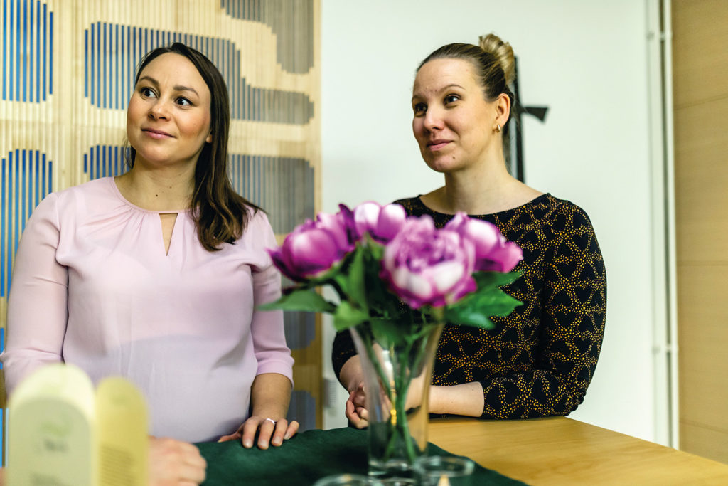 De två kvinnorna står bredvid varandra, på bordet en bukett blommor.
