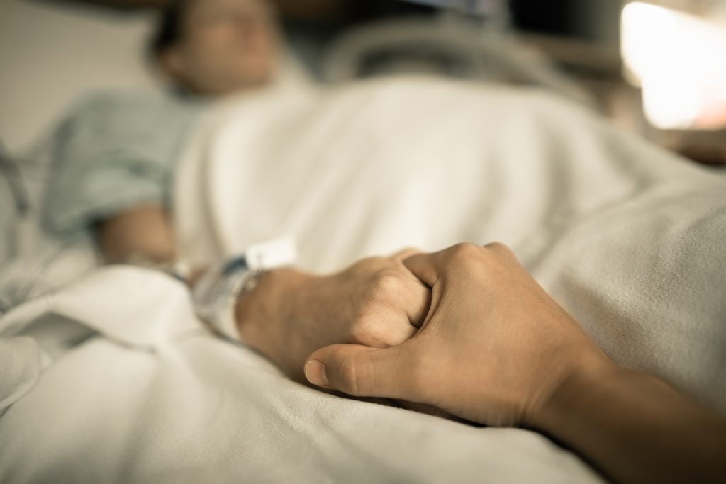 Någon håller handen på en människa som ligger i en sjukhussäng.