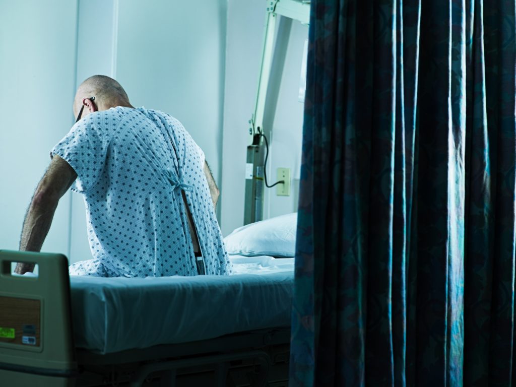 Mies istumassa sairaalan sängyssä sairaalavaatteissa. Kuvattu takaapäin. 
