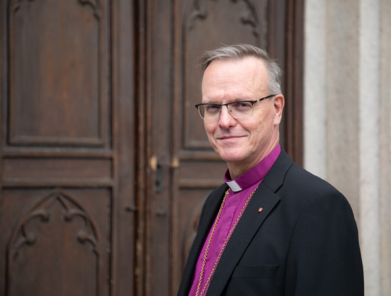 Ärkebiskopen är klädd i lila biskopsskjorta och svart kavaj, han har grått hår och glasögon och står framför en brunmålad kyrkdörr.