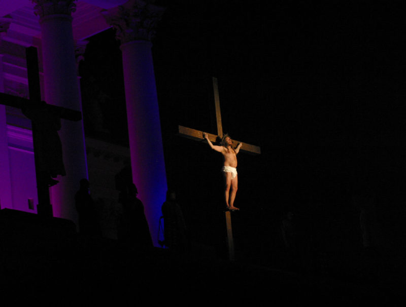Passionsspelets Jesus hänger på korset, upplyst framför en mörk kyrkfasad.