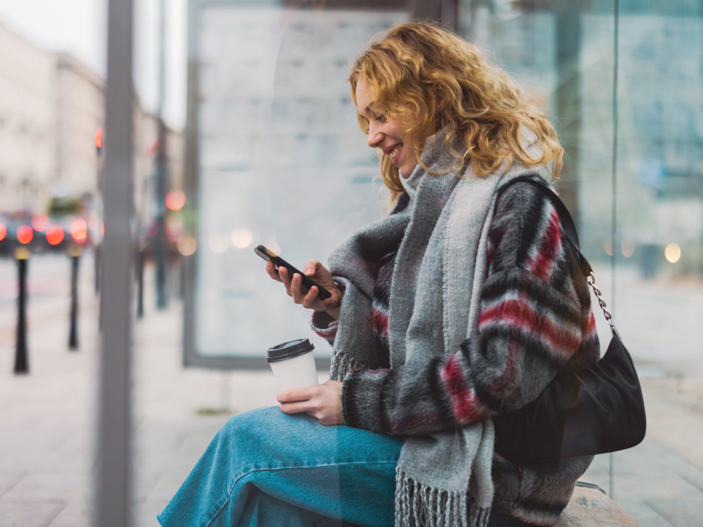 Kvinna sitter vid busshållplats och ler mot något på sin telefon.