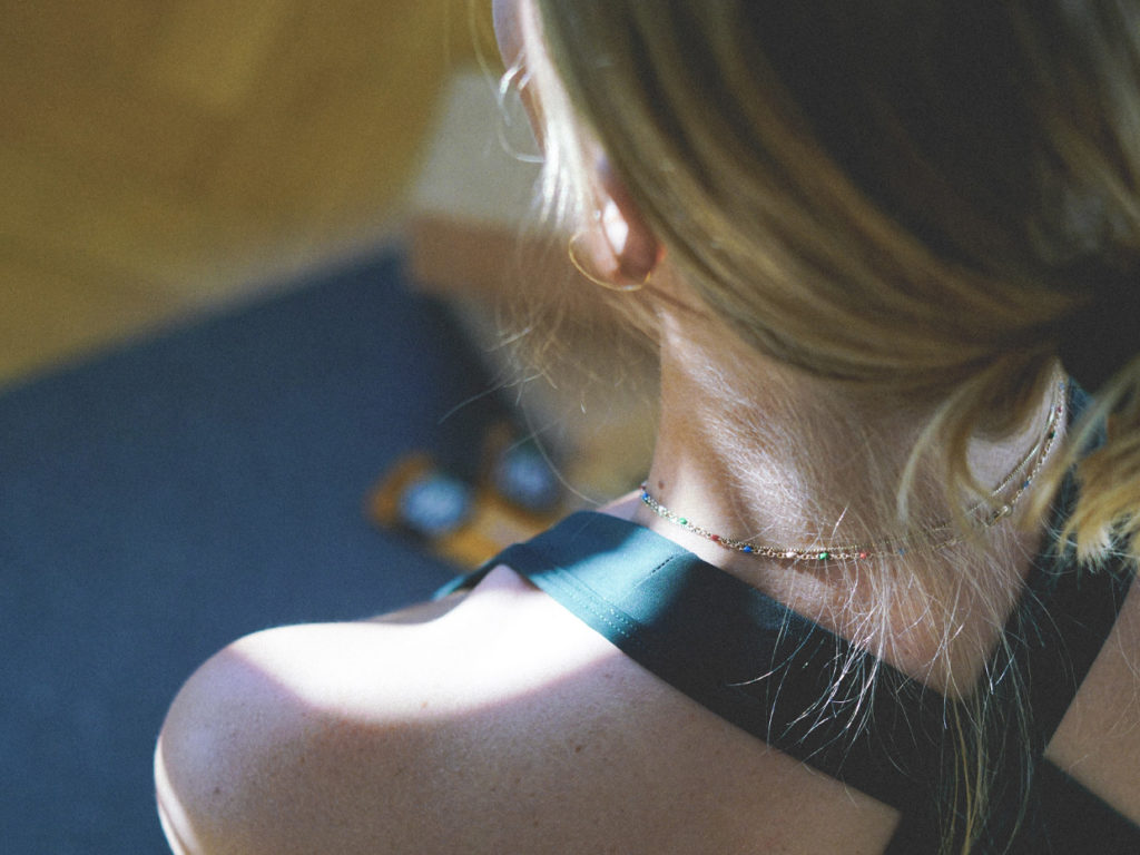 Närbild tagen över axeln på en kvinna som är klädd i sporttopp med bara axlar, har håret uppsatt en låg knut och tunna guldsmycken kring halsen.