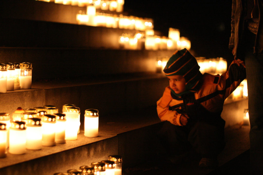 Ett litet barn sitter och tittar på alla gravljus som brinner vid en minnessten på kyrkogården.