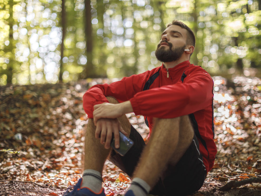 En man i träningskläder sitter på marken i skogen, han blundar med huvudet bakåtlutat och lyssnar på något i sina hörlurar.