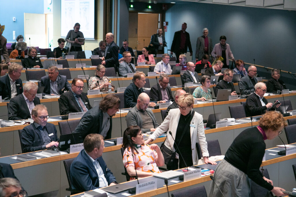 Kyrkomötet ombud sitter i auditorium, en del samtalar, många tittar på sina datorer eller telefoner.