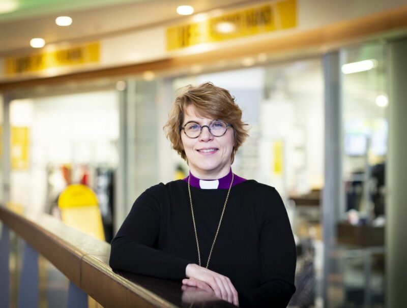 Biskopen är klädd i lila biskopsskjorta och svart tröja, hon har kortklippt, ljust hår och glasögon och står inne i ett köpcenter.