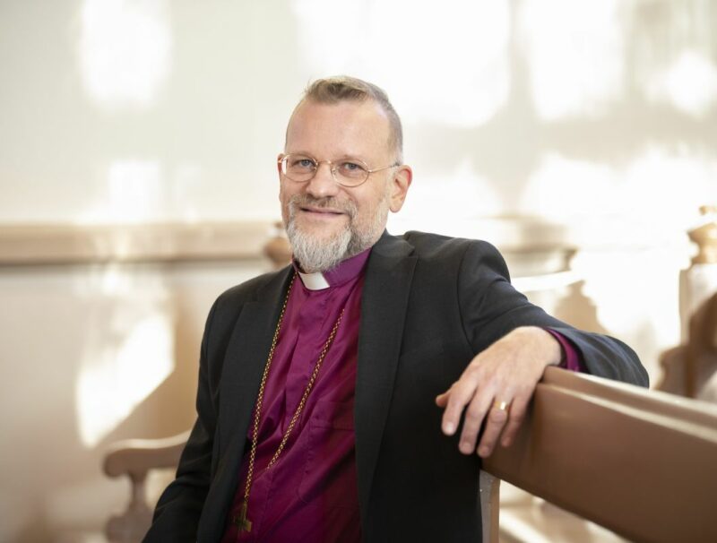 Biskopen är klädd i lila biskopsskjorta och svart kavaj, han har grått hår, skägg och glasögon och sitter i en kyrkbänk.