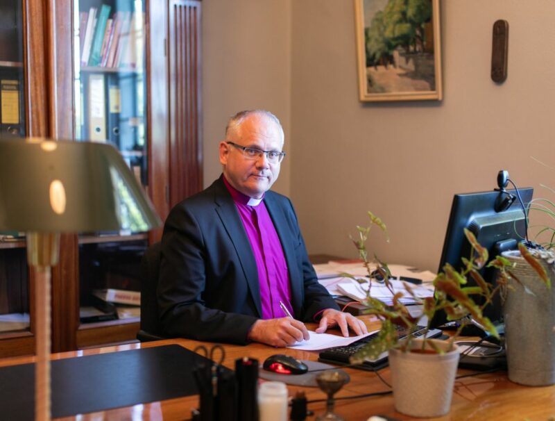 Biskopen är klädd i lila biskopsskjorta och svart kavaj, han har kortklippt, grått hår och glasögon och sitter vid ett stort, gammaldags skrivbord i ett kontorsrum.